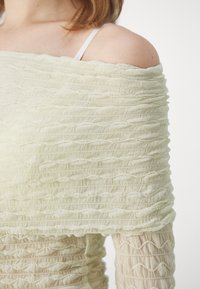 Lys grøn off-the-shoulder top med struktureret, draperet stof. Har et samlet design, blød tekstur og subtile mønstrede accenter.