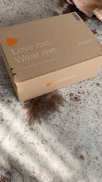 Bruine kartonnen doos met witte tekst "Hou van me. Draag me." en een oranje logo op een gestructureerd, lichtgekleurde ondergrond.