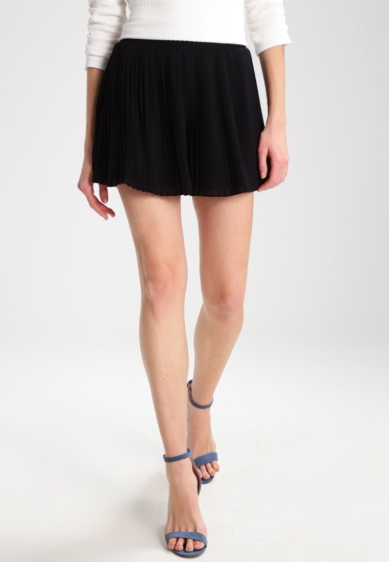 Femme portant une mini-jupe plissée noire, un haut blanc à manches longues et des sandales à talons hauts bleu à bride autour de la cheville, debout sur un fond blanc.