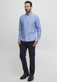 Camicia azzurro chiaro in misto lino, con un piccolo logo e colletto, indossata con pantaloni blu scuro e scarpe nere.