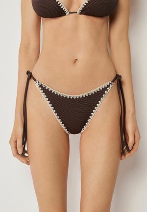 Kvinde iført en mørkebrun bikini med hvid bølget kant, med bindebånd i siden og struktureret stof mod en ensfarvet baggrund.