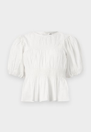 Femme aux cheveux tressés portant un chemisier blanc à manches bouffantes, un short à motifs, des chaussettes marron, des ballerines beige et tenant un petit sac blanc.