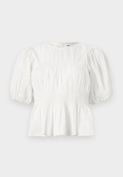 Blouse blanche avec manches trois-quarts bouffantes, corsage froncé et ourlet à basque, présentée sur un fond uni.