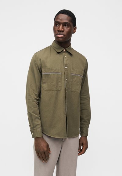 Μακρυμάνικο πουκάμισο σε ελιάς πράσινο χρώμα με κουμπιά μπροστά, δύο τσέπες στο στήθος και αντίθετες ραφές· το ύφασμα φαίνεται λείο και υφασμάτινο.