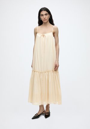 Kvinde med langt mørkt hår iført ærmeløs maxi-kjole i sart ferskenfarve med samlet kant og sorte spidse sko med lav hæl, stående på hvid baggrund.