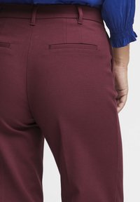 Burgundske prilagojene hlače z gladko teksturo, ki imajo dva zadnja žepa in ozek pas, v kombinaciji z modro bluzo.