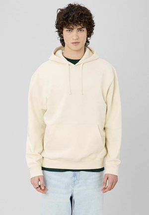 Sweat à capuche couleur crème en tissu doux, avec une poche kangourou, une capuche à cordon de serrage et des poignets côtelés. Porté sur une chemise vert foncé.