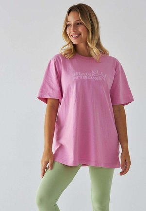 Rosa Baumwoll-T-Shirt mit weißem "Pilates Princess"-Grafik, lässiger Schnitt, kurze Ärmel. Kombiniert mit grünen Leggings, vor einem neutralen Hintergrund.