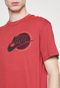 Mies, joka on pukeutunut punaiseen Nike-t-paitaan, jossa on musta logo ja pyöreä teksti rinnassa, leikkattu kaulasta ja hihoista.