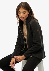 Czarna polarowa kurtka z wysokim kołnierzem, wyposażona w złoty zamek błyskawiczny i brązową naszywkę z logo na rękawie. Gładka struktura i dopasowany krój.