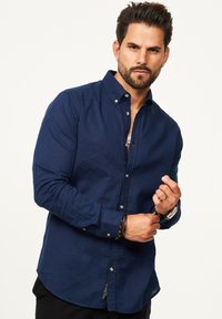 Navy-blaues Langarm-Hemd mit Knopfleiste aus strukturiertem Stoff, mit klassischem Kragen und dezentem Logo am Saum. Die Knöpfe sind in Silberfarbe.
