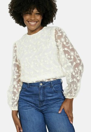 White embroidered long-sleeve blouse with floral patterns, sheer sleeves, and a rounded neckline, paired with high-waisted blue jeans.