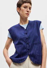 Tamsiai mėlyna be rankovių liemenė su raudonomis juostelėmis, kreiva apatinė dalis ir keturi mygtukai. Dvi priekinių kišenių su lygia apdaila, dėvima virš šviesiai mėlynos marškinės.