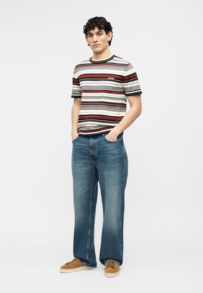 Young man wearing a short-sleeve striped shirt, wide-leg blue jeans, and brown sneakers, standing with hands in pockets.