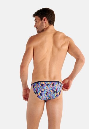 Mann von hinten gezeigt, der bunte florale Badehosen vor einfachem hellem Hintergrund anpasst.