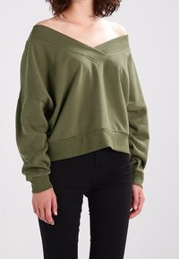 Olivgrön avtagbar sweatshirt med långa ärmar och en avslappnad passform, med en V-formad halsringning och ribbad nederkant. Svarta byxor under.