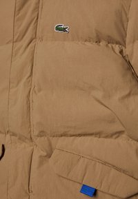 Hnědá péřová bunda s prošívanou texturou, modrým látkovým akcentem a malým zeleno-bílým logem Lacoste na hrudi.