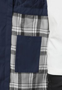 Navy quilted jacket interior with gray and black plaid flannel, featuring a navy patch pocket and smooth texture.