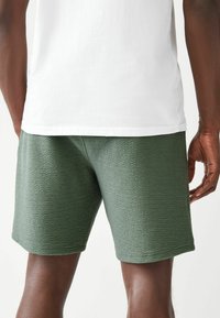 Bermuda verde texturato con un motivo intrecciato, caratterizzato da una vestibilità dritta e un orlo semplice. Indossato con una camicia bianca semplice.