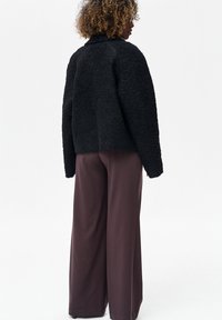 Czarna, teksturowana, miękka kurtka z wysokim kołnierzem, pokazana od tyłu, zestawiona z szerokimi, brązowymi spodniami. Prosty, funkcjonalny design.