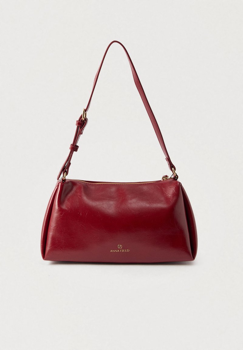 Sac à main en cuir rouge avec un design incurvé, une fermeture éclair supérieure et une sangle ajustable. Présente une texture subtile et un logo de marque embossé à l'avant.