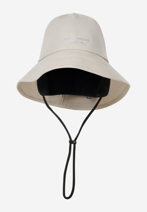Mujer con cabello largo y castaño que lleva un sombrero beige de ala ancha y una chaqueta impermeable verde, ajustando el ala del sombrero con una mano.