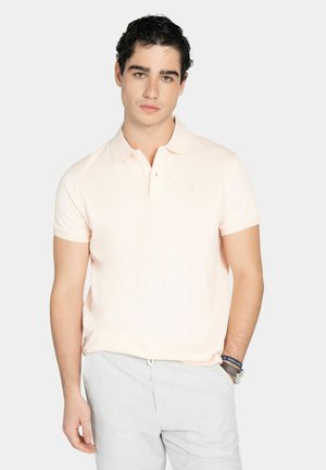 Hombre joven con cabello rizado oscuro, vistiendo una camiseta polo rosa claro y pantalones grises claros a rayas, posando frente a un fondo liso.