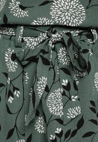 Tessuto verde con finitura testurizzata, caratterizzato da un motivo floreale in bianco e nero. Include una fascia in vita coordinata da annodare per la regolazione.