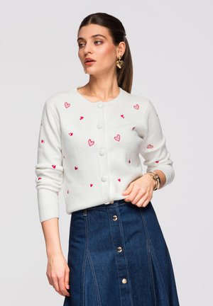 Mujer con suéter blanco abotonado con patrones de corazones rojos y falda de mezclilla azul de talle alto con botones al frente, complementada con joyería dorada.