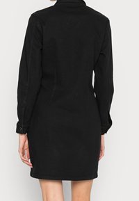 Mustahdin mustasta denim-mekosta, jossa on pitkät hihat, nappi-resorit, kauluksellinen kaula-aukko ja istuva muoto. Sileä rakenne ja polven mittainen helma.