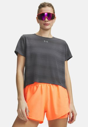 Jonge vrouw die een donkergrijs mesh sportshirt draagt, feloranje sportshorts en reflecterende paarse sportzonnebril tegen een witte achtergrond.