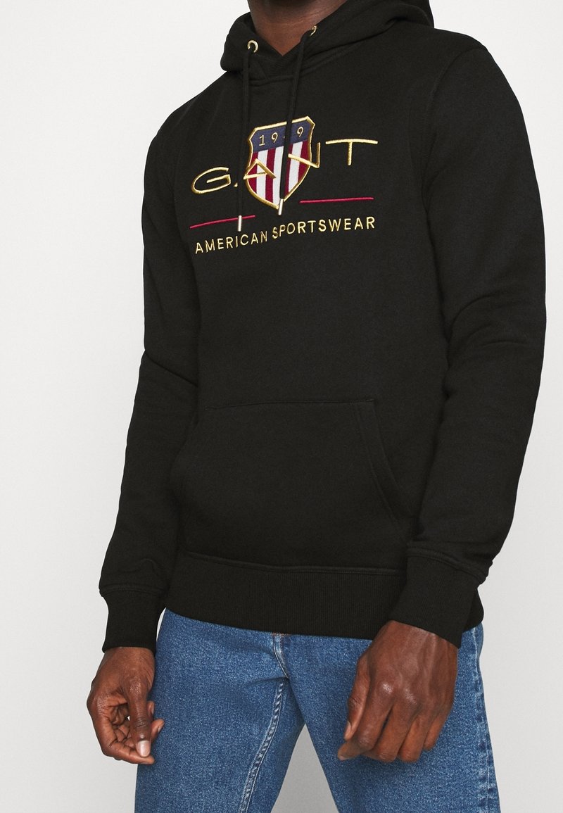 Homme portant un sweat à capuche noir GANT avec un logo en forme de bouclier et le texte « American Sportswear », associé à un jean bleu, montré des épaules aux cuisses.