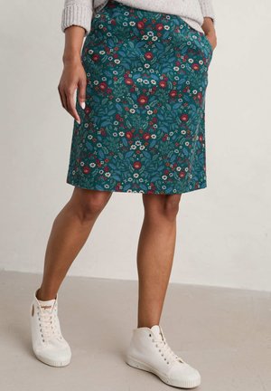 Kobieta w zielono-niebieskiej spódnicy w kwiaty, jasnym swetrze i białych trampkach na wysokiej cholewce stoi z jedną ręką w kieszeni.