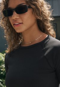Zwart geribd top met lange mouwen en een ronde halslijn, gecombineerd met donkere rechthoekige zonnebrillen en een dunne gouden ketting.