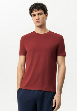 Jonge man met krullend donker haar die een effen rode korte mouw T-shirt en donkerblauwe broek draagt tegen een witte achtergrond.