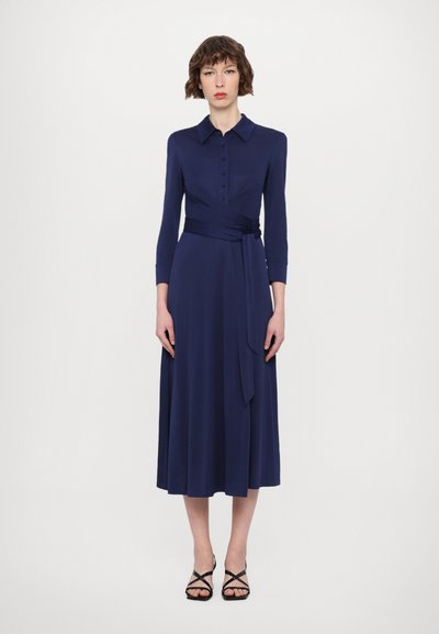 Tamnoplava haljina s ovratnikom, dugim rukavima, kopčanjem na gumbe sprijeda, detaljem omatanja oko struka i lepršavom suknjom do koljena.