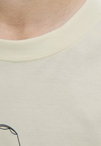 T-shirt en coton beige clair avec un col rond et un motif de dessin linéaire simple près de l'ourlet. Texture lisse, design minimaliste.