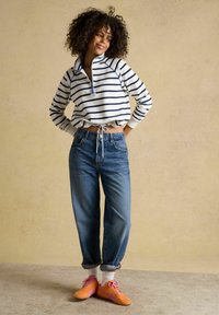 Femeie cu păr creț, purtând un pulover alb cu dungi bleumarin, blugi albaștri, șosete albe și adidași portocalii, stând în fața unui perete bej.