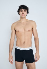 Boxer briefs de algodón negros con un logo blanco de Calvin Klein en la cintura. El modelo está sin camiseta, mostrando una pose neutral.