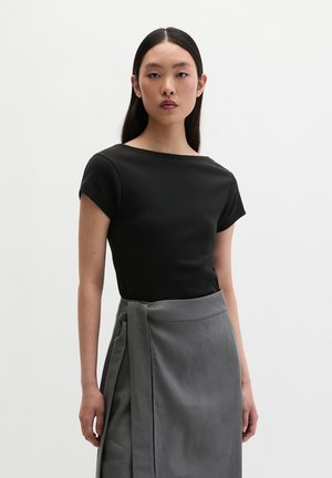 Junge Frau mit langen dunklen Haaren trägt ein schwarzes kurzärmeliges Oberteil und einen hoch taillierten grauen Wickelrock vor einem schlichten weißen Hintergrund.