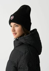 Czarna, żebrowana czapka beanie z okrągłym logo, noszona z czarną kurtką puchową z subtelnym wzorem w panterę i wysokim kołnierzem.