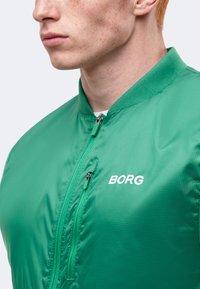 Νέος άνδρας με ανοιχτόχρωμο δέρμα που φοράει πράσινο μπουφάν με φερμουάρ και λογότυπο "BORG" στο στήθος, στέκεται μπροστά σε απλό φόντο.