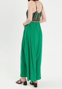 Groene maxi-jurk met een gemusterde lijfje, elastische tailleband en dunne bandjes. De stof is soepel met een vloeiende, relaxte pasvorm. Vierkante haksandalen.