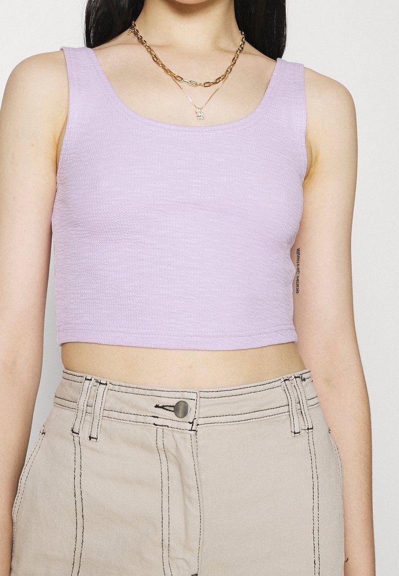 Débardeur court lilas en tissu texturé, avec un col rond et de larges bretelles, associé à un pantalon beige taille haute.