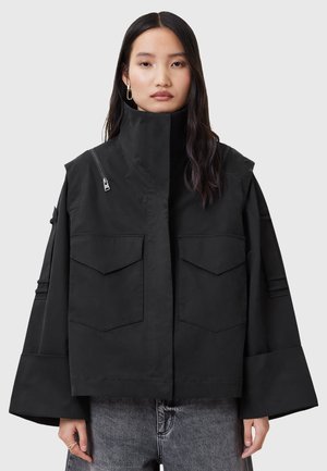 Black cropped jacket with a high collar, large front pockets, and wide sleeves. Features a side zipper and smooth, durable fabric.