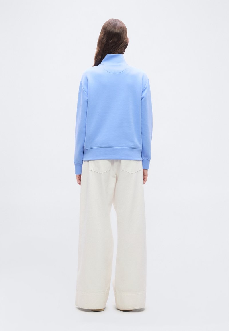 Personne aux longs cheveux bruns portant un haut à manches longues bleu clair et un pantalon blanc à jambes larges, debout, dos à un fond uni.