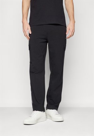Pantalons cargo noirs en tissu doux, dotés de poches latérales, de jambes fuselées et d'un logo discret sur la poche gauche. Portés avec des baskets blanches.