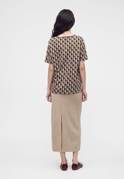 Femme aux longs cheveux foncés portant un haut à manches courtes à motif géométrique, une jupe midi beige avec une fente au dos et des chaussures marron foncé, de dos.