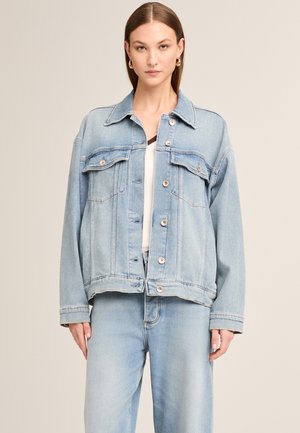 Woman with straight hair wearing gold hoop earrings, light blue oversized denim jacket buttoned halfway, and matching jeans.