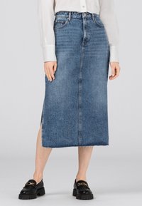 Kvinna iklädd en midilång blå jeanskjol med sidofrånskärning, vit knäppt skjorta och svarta loafers med detaljer i guldkedja.